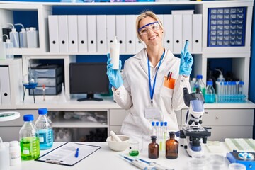 Fototapeta premium Young caucasian woman working at scientist laboratory holding body lotion smiling with an idea or question pointing finger with happy face, number one