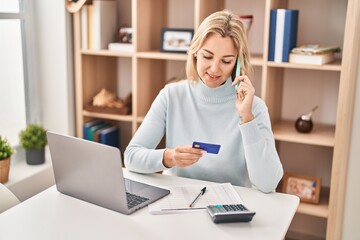 Young blonde woman talking on the smartphone using credit card at home