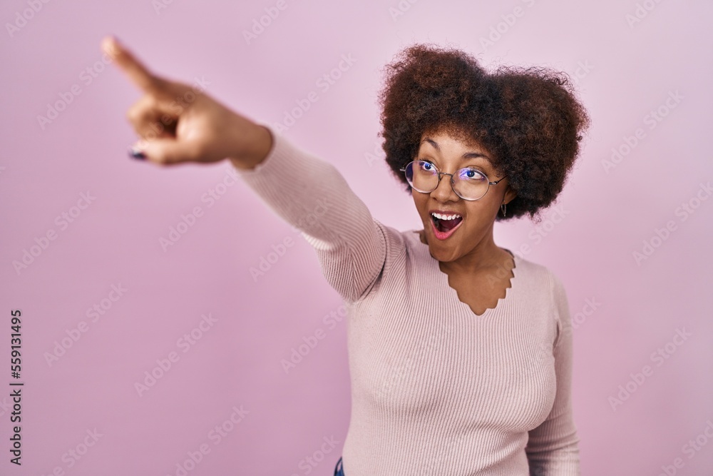 Sticker Young african american woman standing over pink background pointing with finger surprised ahead, open mouth amazed expression, something on the front