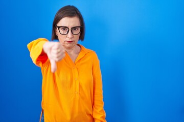 Middle age hispanic woman wearing glasses standing over blue background looking unhappy and angry showing rejection and negative with thumbs down gesture. bad expression.
