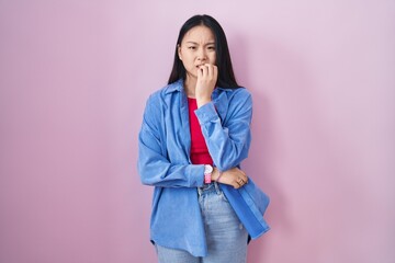 Young asian woman standing over pink background looking stressed and nervous with hands on mouth biting nails. anxiety problem.