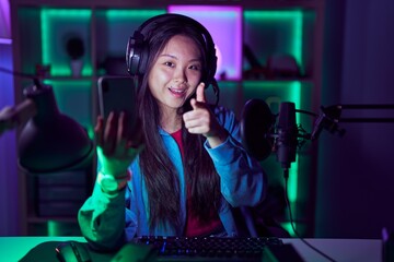 Young asian woman playing video games with smartphone pointing fingers to camera with happy and funny face. good energy and vibes.