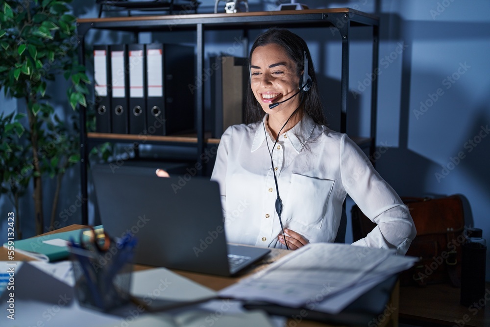 Poster young brunette woman wearing call center agent headset working late at night smiling friendly offeri