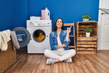 Hispanic woman doing laundry sitting on the floor smiling happy pointing with hand and finger to the side