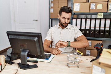 Handsome hispanic man working at small business commerce checking the time on wrist watch, relaxed and confident