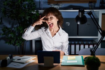 Middle age woman working at the office at night doing peace symbol with fingers over face, smiling...
