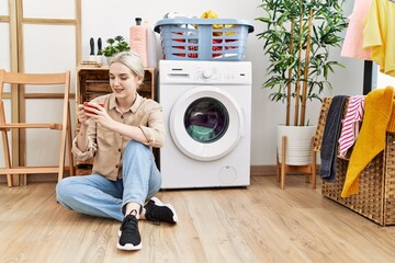 Young caucasian woman drinking coffee waiting for washing machine at laundry room
