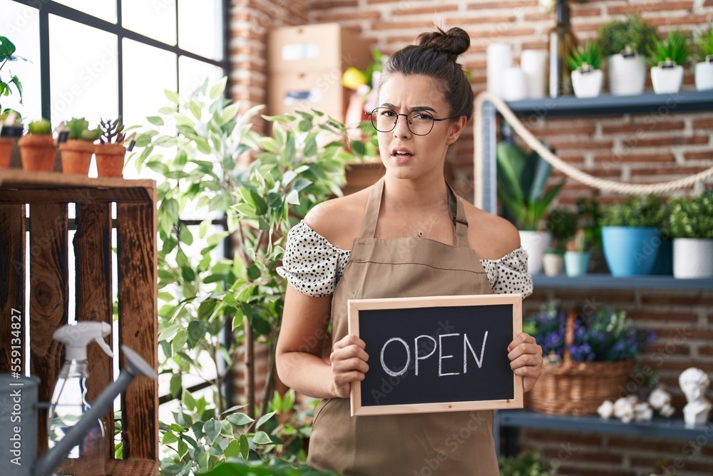 Poster Hispanic woman working at florist holding open sign clueless and confused expression. doubt concept.