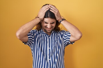 Hispanic man with long hair standing over yellow background suffering from headache desperate and stressed because pain and migraine. hands on head.