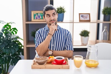 Hispanic man with long hair sitting on the table having breakfast serious face thinking about question with hand on chin, thoughtful about confusing idea
