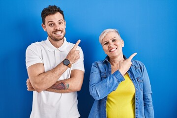 Young brazilian mother and son standing over blue background with a big smile on face, pointing with hand finger to the side looking at the camera.
