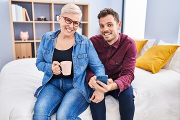 Mother and son using smartphone drinking coffee sitting on bed at bedroom