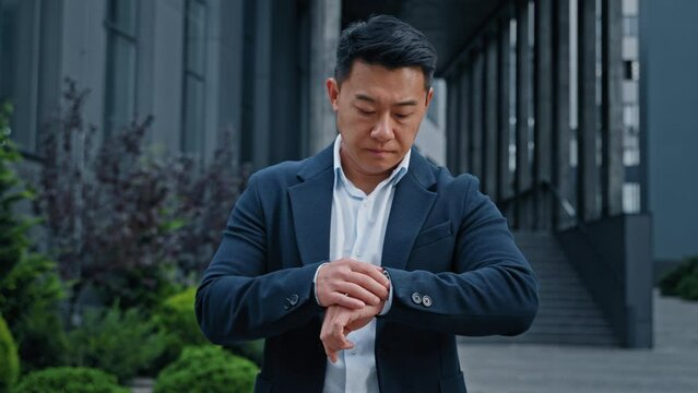 Angry worried middle-aged man 40s Asian Korean businessman standing in city looking at wristwatch check time waiting taxi worry about lateness late time hurry to business meeting inpatient male wait