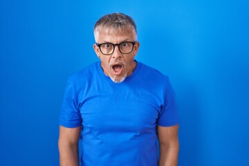 Hispanic man with grey hair standing over blue background afraid and shocked with surprise and amazed expression, fear and excited face.