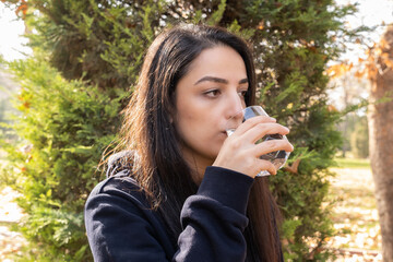 Fototapeta premium Drinking glass of water, thirsty dehydrated woman drinking glass of water. Outdoor, autumn day. Green tree background, copy space. Recovering hydration balance. Healthy habit, lifestyle concept idea.