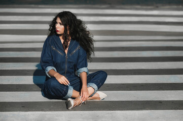 Fototapeta premium A dark-haired girl in denim overalls; a beautiful girl walks along the road through a pedestrian crossing; red lipstick on her lips