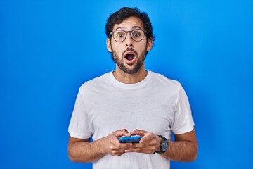 Handsome latin man using smartphone typing message afraid and shocked with surprise and amazed...
