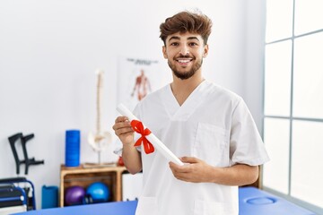 Young arab man wearing physiotherapist uniform holding diploma at clinic