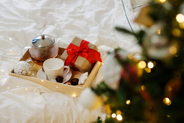 Fototapeta na wymiar Christmas concept - wooden tray with cup, teapot, candies and gift box on the bed near decorated Christmas tree