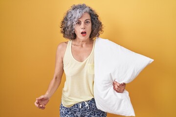 Middle age woman with grey hair wearing pijama hugging pillow in shock face, looking skeptical and...