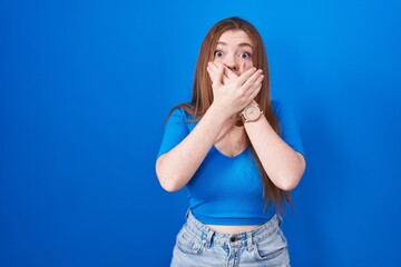 Redhead woman standing over blue background shocked covering mouth with hands for mistake. secret...