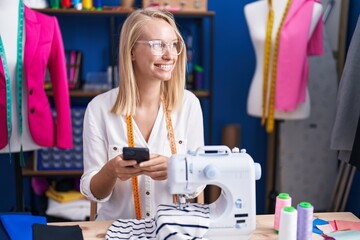 Young blonde woman tailor smiling confident using smartphone at sewing studio