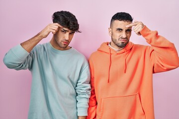 Young hispanic gay couple standing over pink background pointing unhappy to pimple on forehead,...