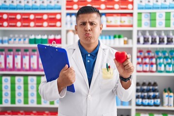 Young hispanic man working at pharmacy drugstore holding heart puffing cheeks with funny face....
