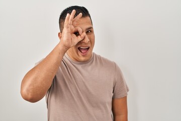 Hispanic young man standing over white background doing ok gesture with hand smiling, eye looking...