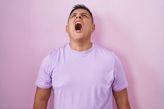 Young hispanic man standing over pink background angry and mad screaming frustrated and furious, shouting with anger. rage and aggressive concept.
