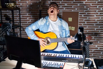 Hispanic young man playing classic guitar at music studio angry and mad screaming frustrated and furious, shouting with anger. rage and aggressive concept.