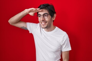 Fototapeta na wymiar Young hispanic man standing over red background very happy and smiling looking far away with hand over head. searching concept.