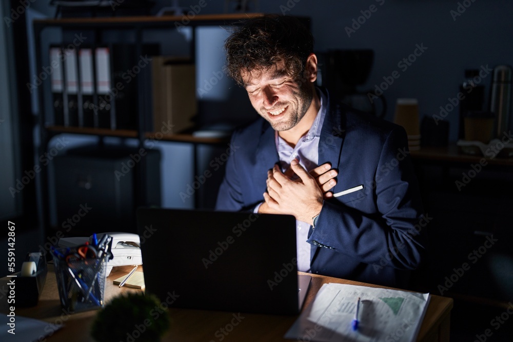 Poster hispanic young man working at the office at night smiling with hands on chest with closed eyes and g