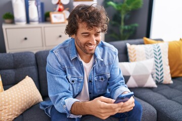 Young hispanic man using smartphone sitting on sofa at home