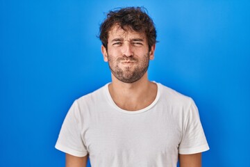 Hispanic young man standing over blue background puffing cheeks with funny face. mouth inflated with air, crazy expression. - obrazy, fototapety, plakaty