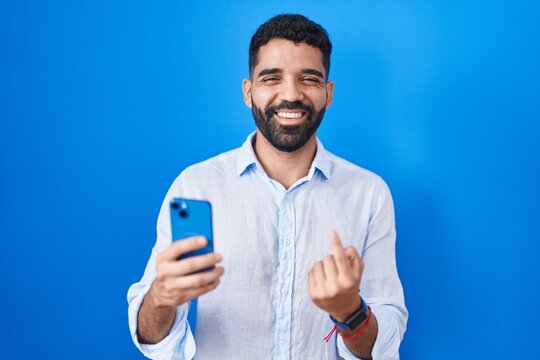 Hispanic man with beard using smartphone typing message beckoning come here gesture with hand inviting welcoming happy and smiling