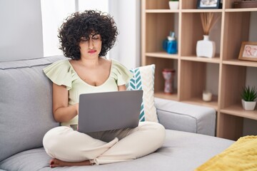 Young brunette woman with curly hair using laptop sitting on the sofa at home depressed and worry for distress, crying angry and afraid. sad expression.