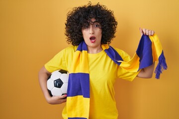 Young brunette woman with curly hair football hooligan holding ball in shock face, looking...