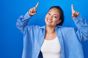 Asian young woman standing over blue background smiling amazed and surprised and pointing up with fingers and raised arms.