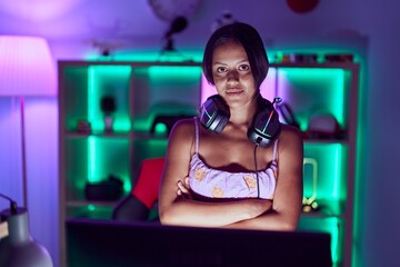 Fototapeta na wymiar Young hispanic woman streamer wearing headphones standing with arms crossed gesture at gaming room