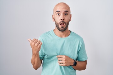 Middle age bald man standing over white background surprised pointing with hand finger to the side, open mouth amazed expression.