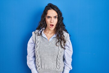 Young brunette woman standing over blue background in shock face, looking skeptical and sarcastic, surprised with open mouth