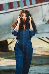 Magnificent volume
curly hair; A dark-haired girl is wearing denim overalls; a sexy girl poses against the background of an airplane; red lipstick on the lips;
