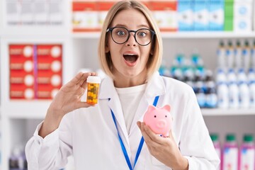 Young caucasian woman working at pharmacy drugstore holding pills an piggy bank celebrating crazy and amazed for success with open eyes screaming excited.