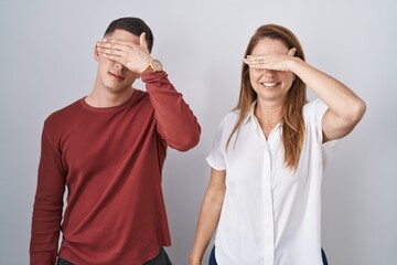 Mother and son standing together over isolated background covering eyes with hand, looking serious and sad. sightless, hiding and rejection concept