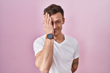 Caucasian man standing over pink background covering one eye with hand, confident smile on face and surprise emotion.