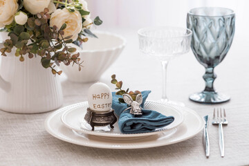 Beautiful spring table setting. A plate with a cotton napkin with a bunny. Silverware and a vase of flowers on a linen tablecloth. The concept of festive serving and a bright Easter holiday.