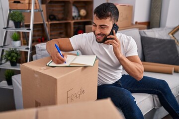 Young hispanic man talking on the smartphone writing on book at new home