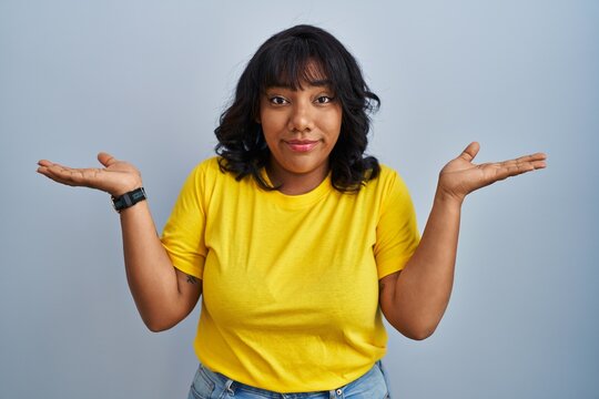 Hispanic woman standing over blue background clueless and confused expression with arms and hands raised. doubt concept.