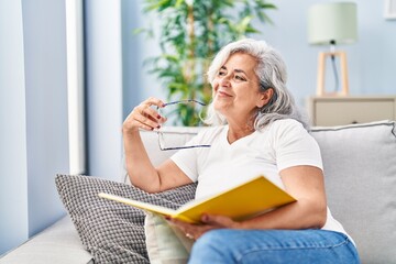 Middle age woman reading book sitting on sofa at home